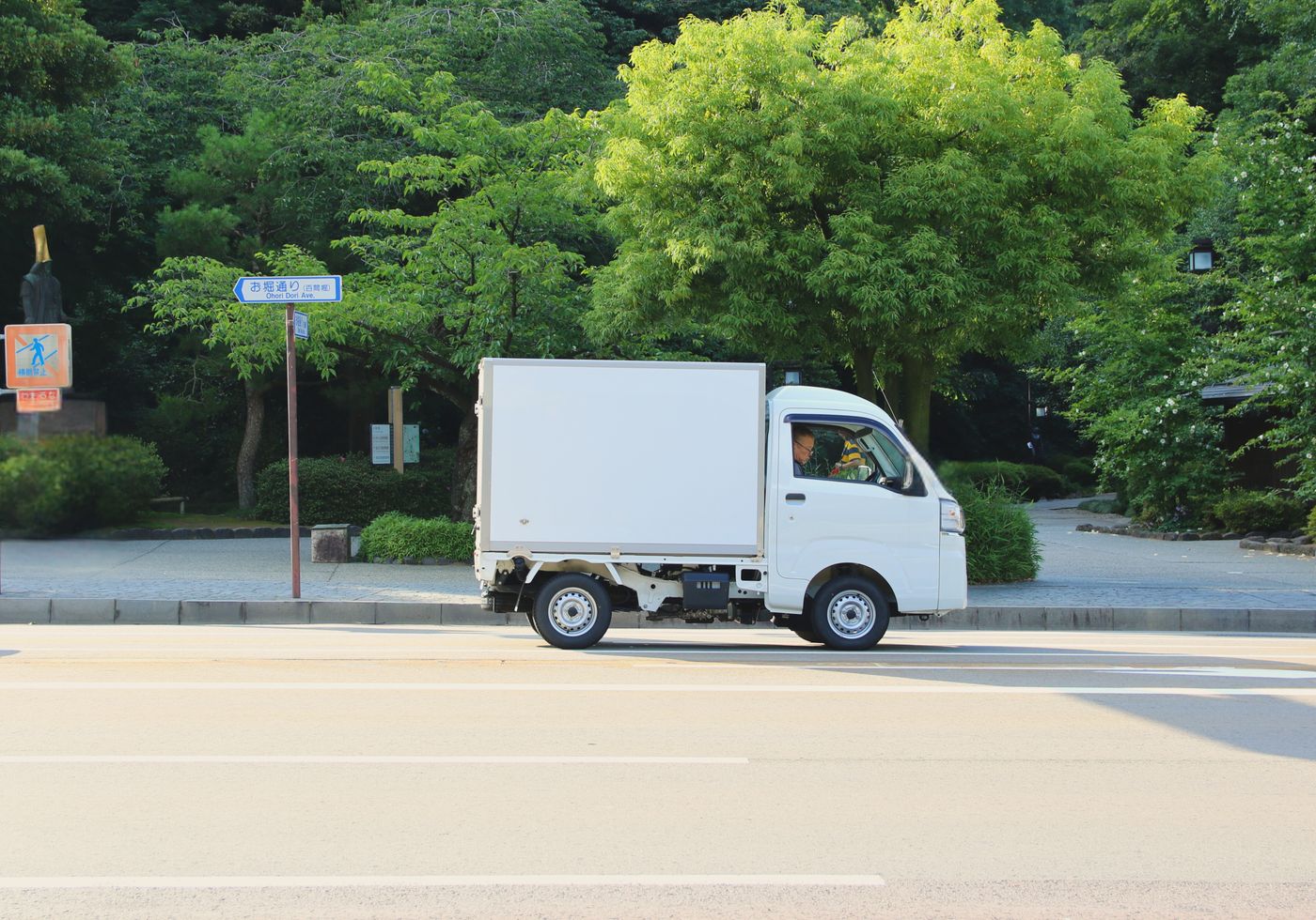 並木のある片側二車線の道を走る白い小型トラック